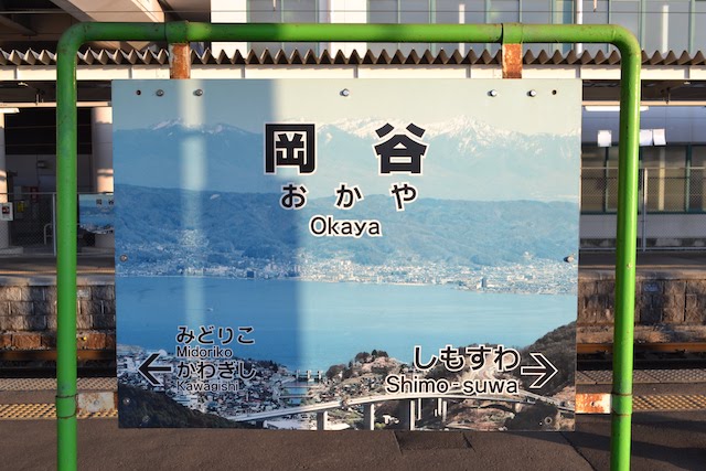 諏訪湖と八ケ岳連峰がデザインされた岡谷駅の写真付き駅名標